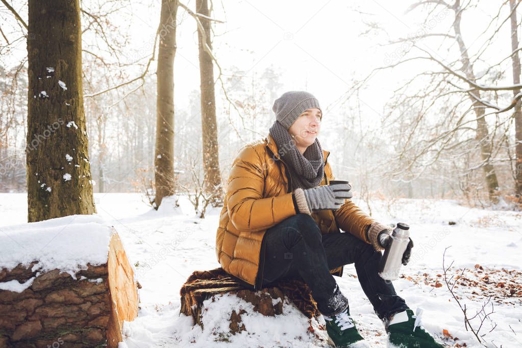 young man holding thermos and sitting in winter forest