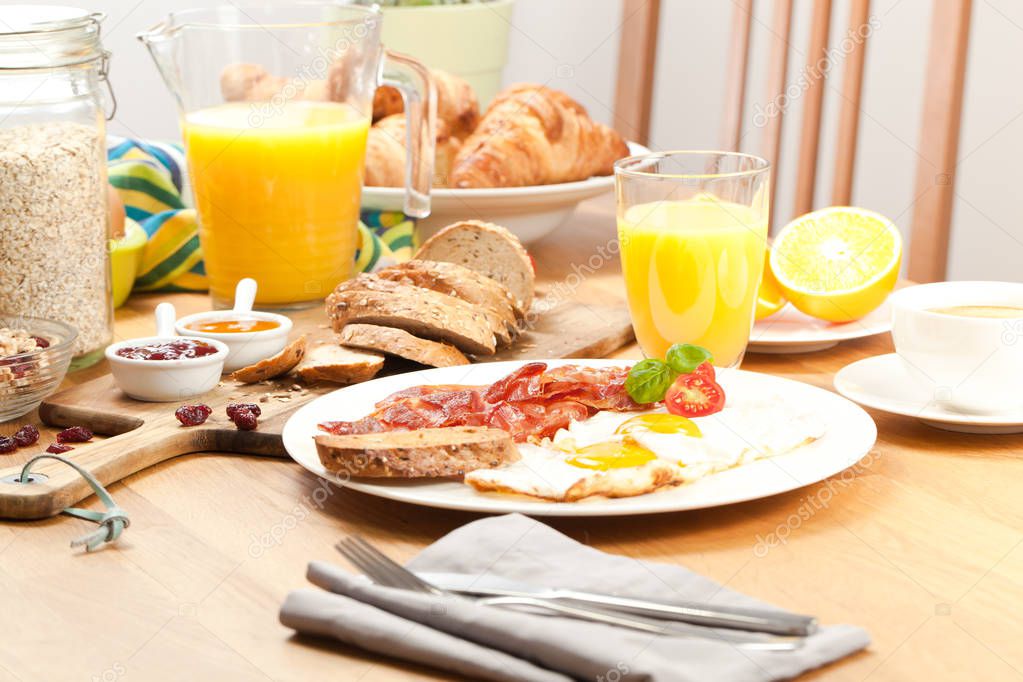 fried eggs with bacon, fork with knife on napkin, orange juice, coffee and croissants for breakfast served on wooden table 