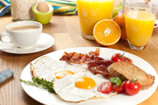 tasty breakfast with fried eggs and bacon on white plate, orange juice, boiled egg, coffee and muesli on wooden table