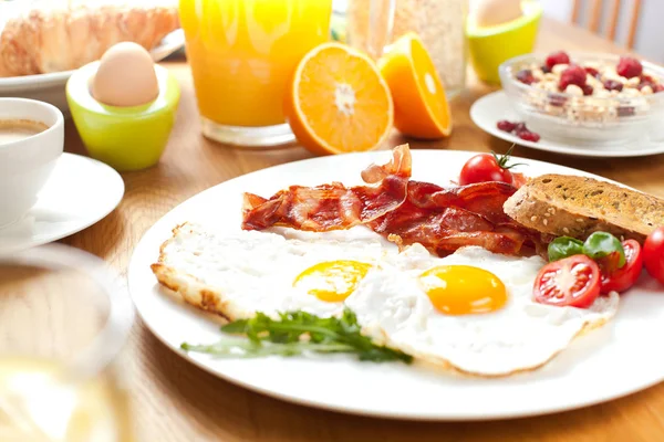 delicious breakfast with fried eggs and bacon, bread, cherry tomatoes, muesli, orange juice and boiled eggs on table