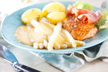 kuşkonmaz, haşlanmış patates, somon steak, marul yaprakları ve mavi plaka üzerinde jambon ile gurme yemek