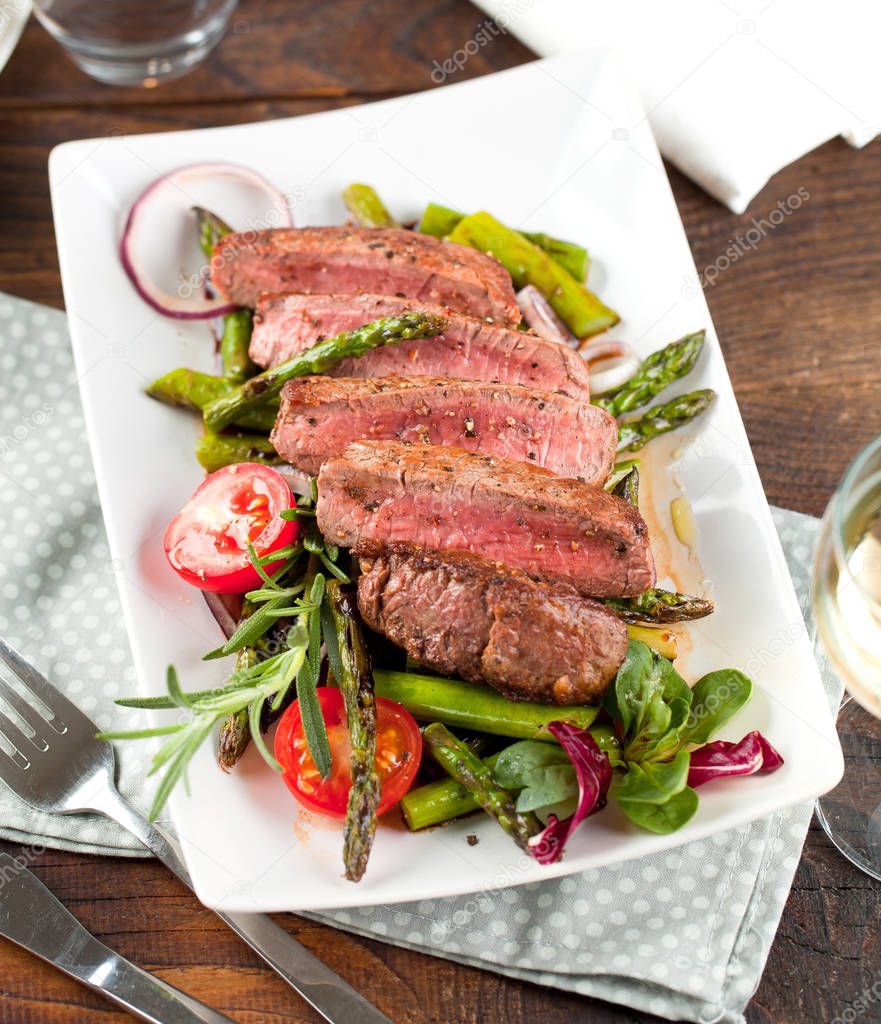 top view of delicious sliced steak with grilled asparagus, tomatoes and herbs on plate 