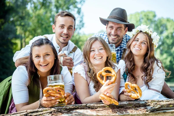プレッツェルでビールのグラスを持ち 屋外のカメラで微笑むバイエルンの衣装で5人の幸せな若い友人 — ストック写真