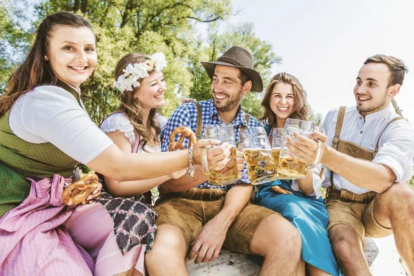 穿着巴伐利亚服装的快乐朋友们在户外喝啤酒和吃椒盐饼干 — 图库照片