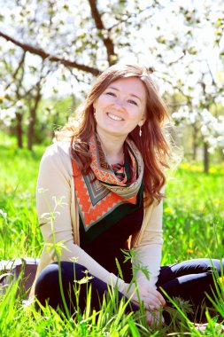 Güzel genç bir kadın yeşil çimlerde oturuyor ve kameraya gülümsüyor.  