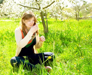 Çiçek açan ağaçların yanında yeşil çimlerde oturan güzel, mutlu genç bir kadın.