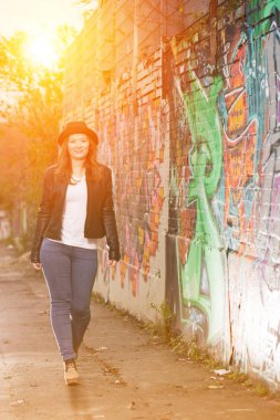Deri ceketli ve şapkalı güzel bir kadın renkli duvar yazılarının arasında yürüyor.