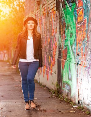 Deri ceketli ve şapkalı güzel bir kadın renkli duvar yazılarının arasında yürüyor.