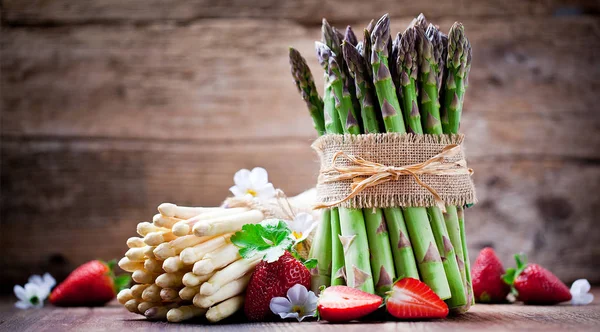 bunches of fresh ripe green and white asparagus, white flowers and red strawberries on wooden table