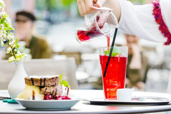 cropped shot of person pouring juice in fresh delicious red ice tea, dessert in cafe on table