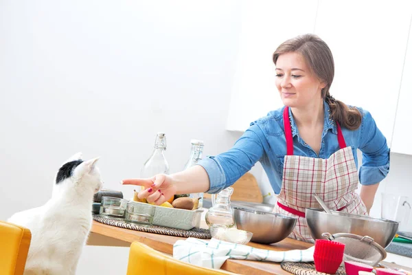 微笑的年轻女子在围裙看着猫 而在厨房做饭 — 图库照片