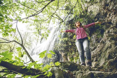 ormanda güzel şelale yakınında açık kollarla duran sırt çantası ile gülümseyen genç kadın düşük açı görünümü