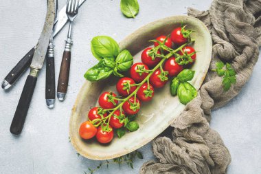 taze olgun kiraz domates üst görünümü, tabakta fesleğen yaprakları ve gri yüzeyde çatal 