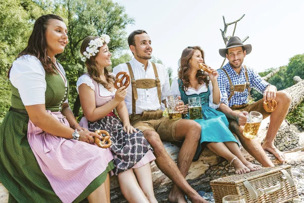 坐在户外时 穿着巴伐利亚服饰的快乐年轻朋友拿着啤酒和薄饼杯的低角度视角 — 图库照片