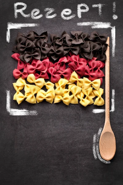 top view of uncooked pasta in colors of german flag, wooden spoon and word rezept on black background
