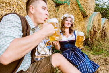 bavyera kostümleri mutlu genç çift saman balyaları yakınında oturan ve bira içme, oktoberfest kavramı