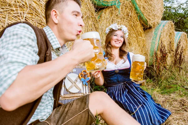ストローベールの近くに座ってビールを飲むバイエルンの衣装で幸せな若いカップル オクトーバーフェストコンセプト — ストック写真
