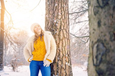 Güzel, gülümseyen genç bir kadın kış ormanında ağacın yanında duruyor.