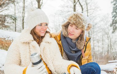 Mutlu genç çift sıcak bir içecek içip kış ormanında birlikte oturuyorlar.