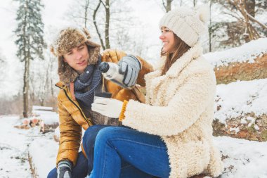 Mutlu genç çift sıcak bir içecek içip kış ormanında birlikte oturuyorlar.