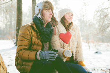 Genç çift ellerinde kırmızı bir kalp ve sıcak bir içecek tutarken birlikte kış parkında oturuyorlar. 