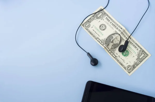Money banknote with black headset and black mobile phone placed on desk with pastel light blue background. Flat lay, space for text. Sound, music and money concept.