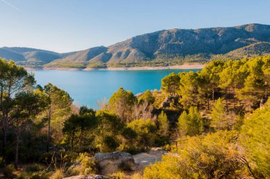 Dağların eteklerinde göl ve güneşli bir gün ağaçlar ile çevrilidir. Ormanda bir yol görebilirsiniz.