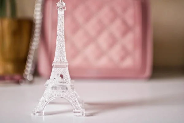 Plastic mini model of Eiffel Tower on white table and biege female bag on blurred background. Travel concept. — Stock Photo, Image