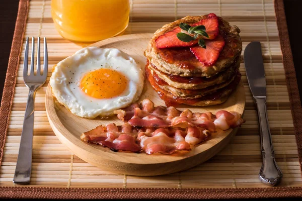 American breakfast consisting of an egg, a few slices of bacon and a pancake of strawberry jam and strawberries. Drink a natural orange juice.