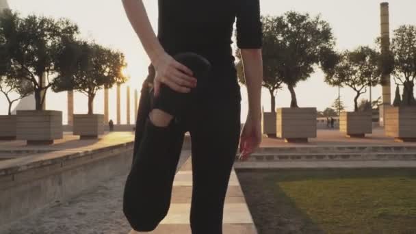 Молода жінка в чорному костюмі розтягує м'язи ніг, перш ніж бігти по вулиці в сонячну теплу погоду. Молода дівчина робить корисні вправи для здоров'я. Жінка згинає ногу назад, щоб накачати — стокове відео