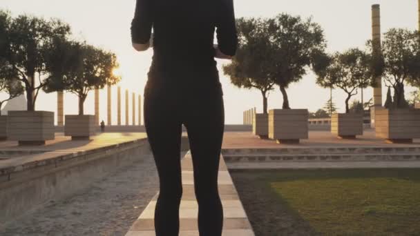 Siyah giyimli genç bir kadın şehir parkında durmuş, koşudan sonra dinleniyor. Koşmadan önce bacaklarına masaj yapıyor. Kız Park'taki patikada koşuyor. Dışarıdaki hava uygun Loya egzersizleridir — Stok video