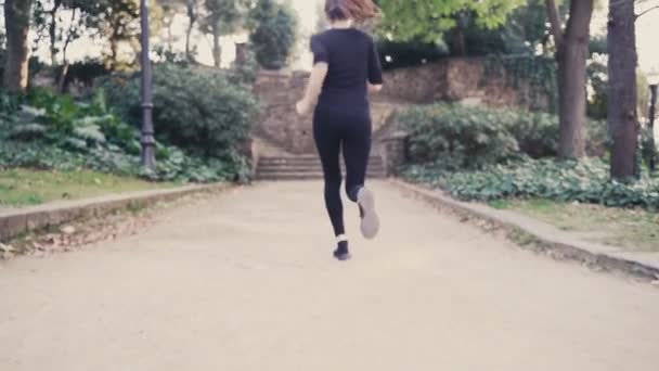 Jonge vrouw in een zwart trainingspak loopt de trap op in het park. Meisje op de vlucht. Het weer is zonnig en warm. Een jong meisje rent de trap op tijdens een hardlooptraining. — Stockvideo