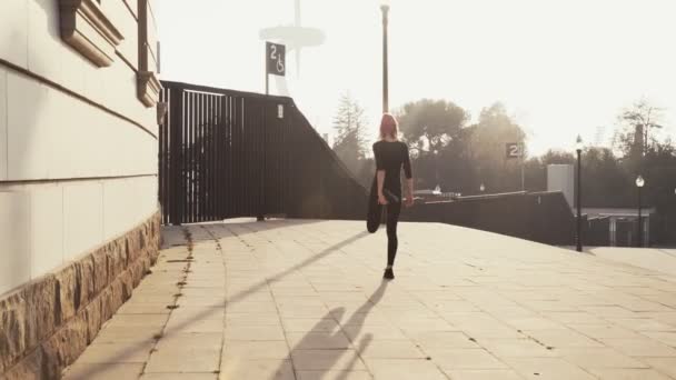 Kvinnan i svart träningsoverall sträcker på benmusklerna innan hon joggar nerför gatan i soligt varmt väder. Ung flicka gör användbara övningar för hälsa och kropp. Kvinnan böjer sitt ben bakåt för att pumpa — Stockvideo