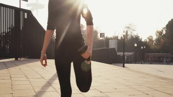 Mädchen im schwarzen Trainingsanzug dehnt Beinmuskeln, bevor sie bei sonnigem, warmem Wetter die Straße hinunterjoggt. junges Mädchen, das nützliche Übungen für die Gesundheit macht. Frau beugt sein Bein nach hinten, um die Muskeln zu pumpen — Stockvideo
