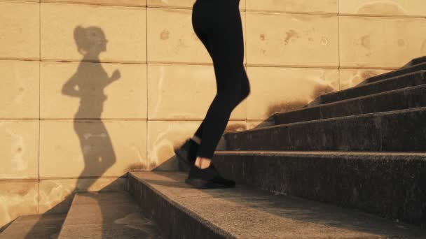 Jonge vrouw in een zwarte trainingspak het runnen van de stappen. Meisje op de vlucht. Het weer is zonnig en warm. Een jong meisje loopt de trap op tijdens een running workout. — Stockvideo