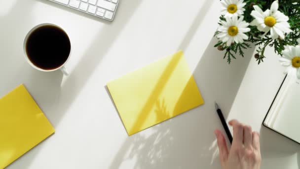Νωρίς το πρωί της άνοιξης στο γραφείο μια νεαρή γυναίκα υπογράφει έναν κίτρινο φάκελο με ένα γράμμα. Το κορίτσι υπογράφει τους φακέλους στους φίλους και τους συναδέλφους του. αρκετούς κίτρινους φακέλους, στο τραπέζι — Αρχείο Βίντεο