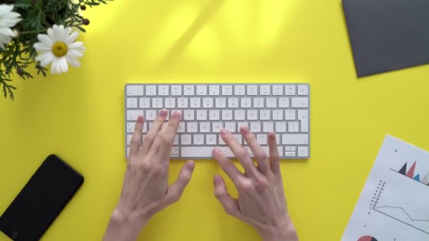 Νεαρή γυναίκα δακτυλογραφεί σε ένα πληκτρολόγιο σε ένα κίτρινο τραπέζι που περιβάλλεται από έγγραφα γραφείου και λουλούδια. Κορίτσι πληκτρολογεί σε ένα σύντομο, μικρό πληκτρολόγιο. Κορίτσι πληκτρολογώντας ένα γράμμα στον υπολογιστή σας σε μια ηλιόλουστη μέρα — Αρχείο Βίντεο