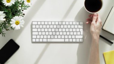 Kadın klavyede yazarken bir fincan kahve getiriliyor. Ofis kağıtları ve çiçeklerle çevrili sarı bir masada klavyeye yazı yazan genç bir kadın..