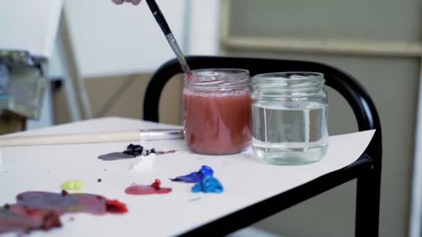L'artista inumidisce un piccolo pennello in un contenitore d'acqua. mescola colori diversi. L'artista mescola verde e blu per ottenere il colore desiderato . — Video Stock