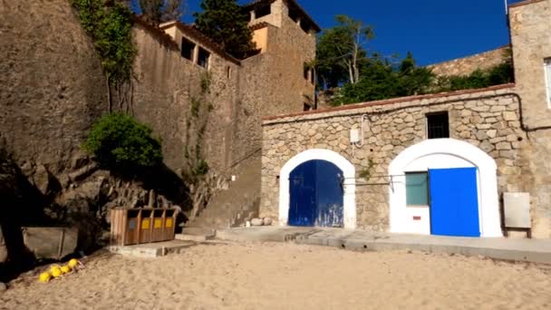 Panorama d'une belle plage de sable avec des garages pour bateaux. Journée ensoleillée dans le village méditerranéen. Sable doré et ciel bleu. Bâtiment en pierre avec portes bleues. Ville côtière méditerranéenne — Video