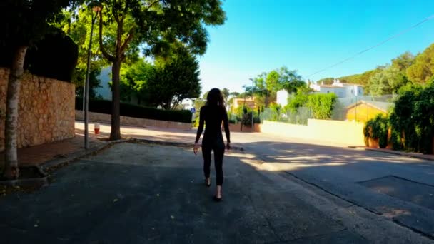 Chica en un traje de neopreno negro caminando por la calle hacia el mar. Día soleado en el pueblo. Triatlón. Listo para nadar. Nadando en aguas abiertas. Nadar por la mañana. Ciudad costera mediterránea — Vídeos de Stock