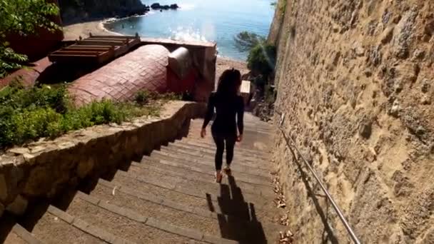 Ένα κορίτσι με μαύρο κοστούμι από νεοπρένιο κατεβαίνει τις πέτρινες σκάλες για τη θάλασσα. Ηλιόλουστη μέρα στο χωριό. Τρίαθλο. Έτοιμοι για κολύμπι. Κολύμπι σε ανοιχτή θάλασσα. Πρωινή κολυμβητική δραστηριότητα. Παράκτιες μεσογειακές πόλεις — Αρχείο Βίντεο