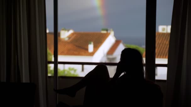 大きな窓の背景に電話で話している少女のシルエット。窓には雨の空と虹の風景。女がソファに座って電話で話してる. — ストック動画