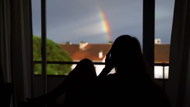Силуэт девушки, разговаривающей по телефону на фоне большого окна. Пейзаж с дождливым небом и радугой в окне. Женщина сидит на диване и болтает по телефону . — стоковое видео