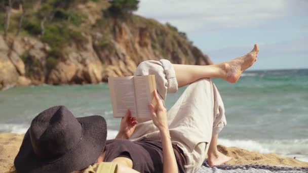Am Strand. Ein Mädchen mit schwarzem Hut liegt am Strand und liest ein Buch. Leichter Wind. Im Hintergrund türkisfarbene Wellen. Sommerkälte. Urlaub am Meer. Ruhe und Vergnügen. Urlaub am Strand. — Stockvideo