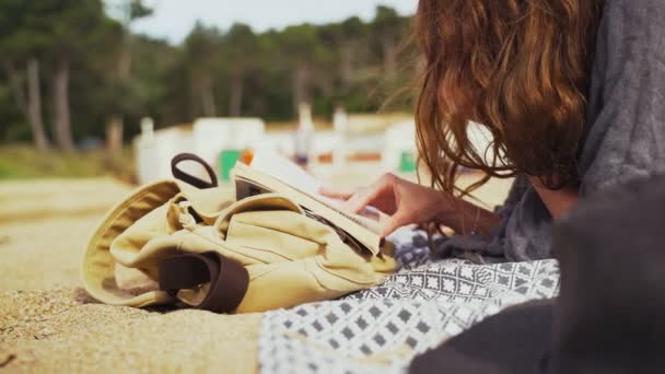 Na praia. Uma ruiva jaz na praia e lê um livro. Brisa leve. Frio de verão. Férias no mar. Descanso e prazer. Férias na praia. Areia e pinheiros no fundo . — Vídeo de Stock