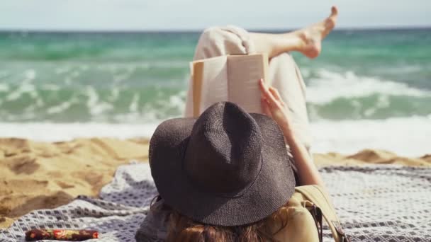 Am Strand. Ein Mädchen mit schwarzem Hut liegt am Strand und liest ein Buch. Leichter Wind. Im Hintergrund türkisfarbene Wellen. Sommerkälte. Urlaub am Meer. Ruhe und Vergnügen. Urlaub am Strand. — Stockvideo