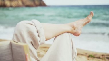 Sahilde. Bir kız plajda uzanıp kitap okuyor. Deniz zemininde kadın bacakları. Hafif bir esinti. Turkuaz dalgalar. Yaz soğuğu. Denizde bir tatil. Dinlen ve keyfine bak. Plajda tatiller.