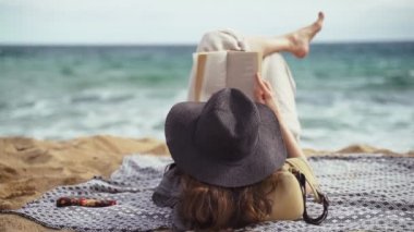 Sahilde. Siyah şapkalı bir kız plajda uzanıp kitap okuyor. Hafif bir esinti. Arka planda turkuaz dalgalar var. Yaz soğuğu. Denizde bir tatil. Dinlen ve keyfine bak. Plajda tatiller.