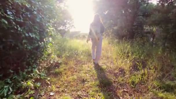 Pěší turistika. Dívka s batohem stojí na lesní stezce a má krásný západ slunce. Zlaté světlo v zeleném lese. Lesní túra. Slunečný letní den. Procházím se mezi stromy a květinami. Dovolená. — Stock video